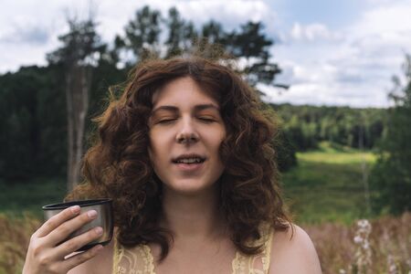 Beautiful young curly-haired red-haired girl with a mug from a thermos on a background of forest and nature in the morningの写真素材