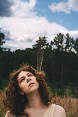 Beautiful young curly-haired red-haired girl on a background of forest and nature in the morningの写真素材