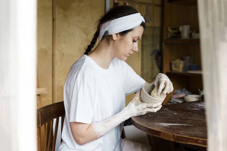 Beautiful young woman in a creative workshop sits at a table and wipes a clay bowl with a sponge. Shaped method for making clay dishes. Handwork. Pottery makingの写真素材