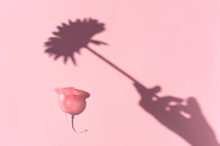 Pink menstrual cup and shadow of a female hand holding a flower on a pink background. Womens Health and Zero Waste Concept. Minimalismの写真素材