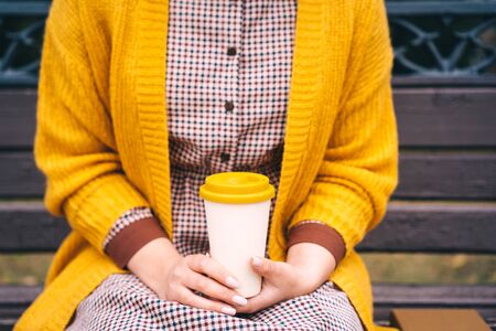 A woman is sitting on a park bench and holding a bamboo cup with hot coffee in her hands. Fashionable autumn clothes in orange mustard tonesの写真素材