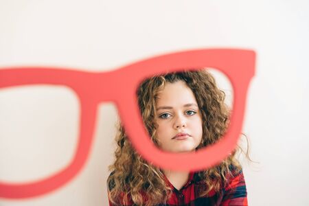 Cute curly long-haired blonde teen girl looks over funny big glasses red color. The concept of interest and good vision.の写真素材