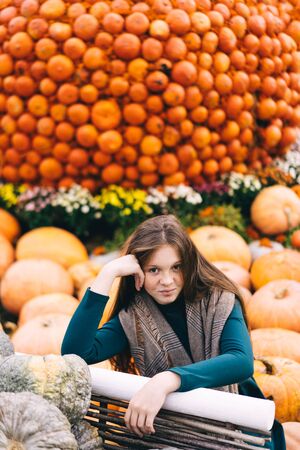 A beautiful red-haired teenage girl with freckles on a pretty little face sits near a cart with pumpkins at the harvesting festival dedicated to the harvest and the end of the summer.の写真素材