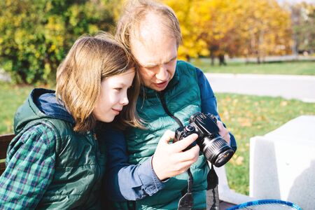 A father and son of school age are talking in close proximity with photo camera sitting on bench. Autumn landscape in yellow tones, warm vests and shirts. Sunny day. Parenthood and childhood conceptの写真素材