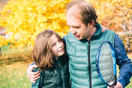 A father and son of school age are talking in close proximity with badminton racket. Autumn landscape in yellow tones, warm vests and shirts. Sunny day. Parenthood and childhood conceptの写真素材