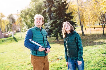 A father and son of school age with badminton racket are looking at sky and talking. Autumn landscape in yellow tones, warm vests and shirts. Sunny funny day. Parenthood and childhood conceptの写真素材