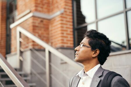 Young handsome Indian man in a white shirt and a business suit. Standing on the street in the city business center near a summer cafe and looking. Break time outdoor office workの写真素材