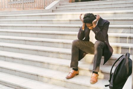 Young handsome Indian man in a white shirt and a business jacket. Sitting sad on the steps of the...
