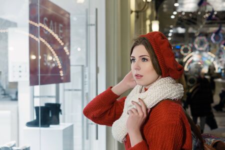 Young beautiful woman in winter beret, white scarf and red knitted warm sweater looking at shop...