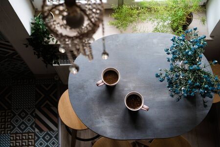 Two ceramic white cups of coffee standing on a wooden oval table by the window in the kitchen....