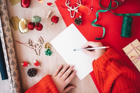 A child in red pullover writing letter to Santa Claus asking for gifts. On the table props for decorating house and the tree on craft paper, clothespins in the shape of hearts for the Advent calendarの写真素材