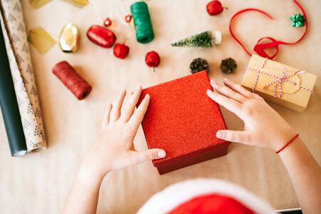 A child in red hat opening red gift box closing. On the table, props for decorating the house and the tree on craft paper, clothespins in the shape of hearts for the Advent calendarの写真素材