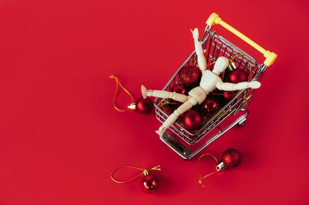 Figurine of a wooden man in a basket for shopping filled with red Christmas decorative balls. Environmental protection concept eco friendly. New year preparation commotionand sale shopping discountの写真素材