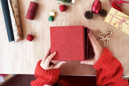 A child hands in red pullover opening red gift box closing. On the table props for decorating house and tree on kraft paper thread, wooden deer head, glitter apple. Creative new year handmade conceptの写真素材