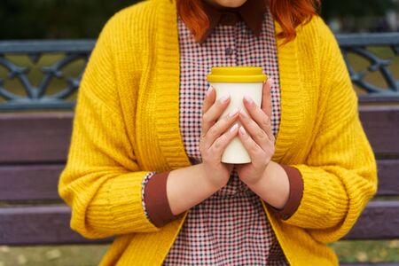 A woman is sitting on a park bench and holding a bamboo cup with hot coffee in hands. Fashionable autumn clothes in orange and yellow warmth mustard tones mood. Pink manicure. Zero waste conceptの写真素材