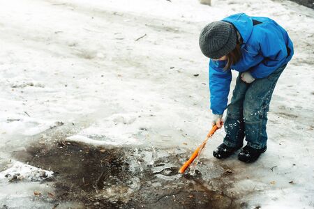 A nine year old school age boy standing in the street in a blue jacket and hat breaking by stick ice in a frozen puddle. Winter frosty sunset of the day walkingの写真素材