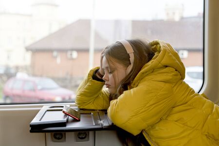 Teenager girl in a yellow jacket with headphones on her head sitting in a suburban train watching a video on a mobile phone. Gadget addictionの写真素材
