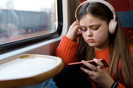 Teenager sad girl 12 years old in red jacket with headphones on her head sitting in suburban train watching a video on a mobile phone. Gadget addiction concept. Spring vacation. Daily trips to schoolの写真素材