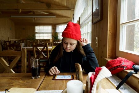 Teenager girl 12 years old in red hat sitting in cafe and playing with phone mobile game app. Gadget addiction concept. Winter vacation Christmas holiday with familyの写真素材