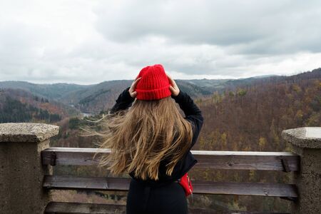 Long-haired girl in a black jacket, a red knitted hat standing on top of a high tower in the...
