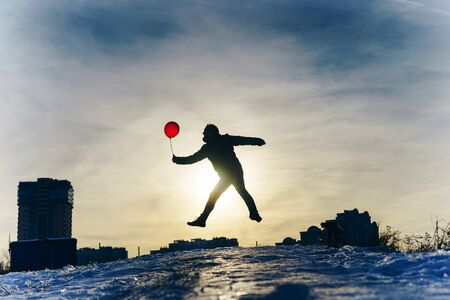 Man from the back in a warm winter jacket of blue color and jeans with a red air rubber balloon in hand against the blue sky on a frosty day. T-shaped silhouette symbolizing freedom and flightの写真素材