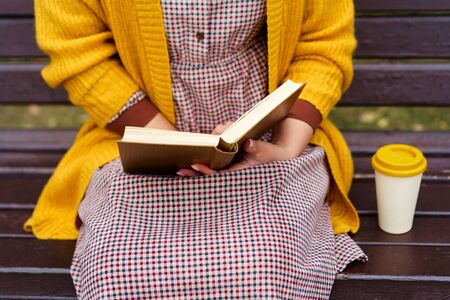 A woman is sitting on a park bench and holding a book and bamboo cup with hot coffee. Fashionable...