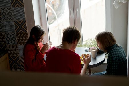 Family breakfast at the oval table by the window. Two boys and girl eating potatoes and drinking coffee from white ceramic mugs. Top view through a crystal chandelier. Weekend Family Customs Conceptの写真素材