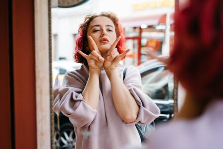 A young woman with red red curly hair makes a face in the door mirror on the street. Funny silly youth conceptの写真素材
