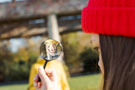 Girl in a red knitted hat looking through a magnifying glass holding glass in her hand on a boy. The concept of exaggeration and distortion of reality at a young ageの写真素材