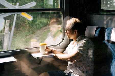 A smiling man in a suburban train looking out the train window at the landscape and holding a bamboo glass for coffee in his hand. Tourist travel concept. Solo travelerの写真素材