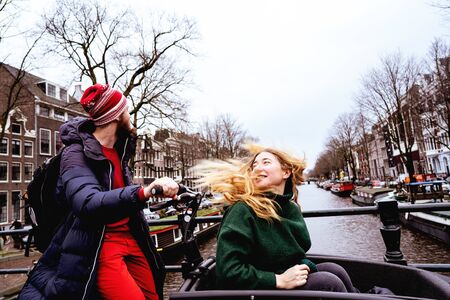 Loving couple man and woman having fun on the streets of Amsterdam in winter warm clothes. Husband rolling wife in the cradle of childrens bike. Funny moments of life together. Valentine day conceptの写真素材