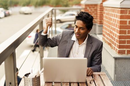 Young handsome Indian man in a white shirt and a business suit is sitting at the table in a summer...