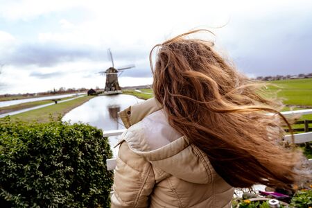 Girl with long red hair developing in the wind. Woman looking into the distance at a typical Dutch provincial landscape with a mill and a canal dividing the fieldsの写真素材