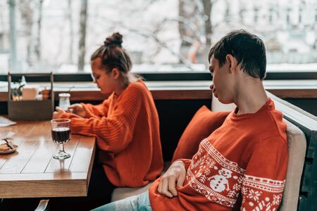 Girl and boy teenager sitting in cafe at a table in red knitted sweater waiting and looking typing in phone messages. Milk chocolate cocktail and the grape juice. Brother and sister at the weekendの写真素材