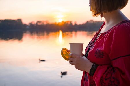 Short haired woman is walking in the park near the river and holding a bamboo cup silicone lid with hot coffee in her hand. Fashionable summer clothes. Beautiful sunset and ducks floating in the pondの写真素材