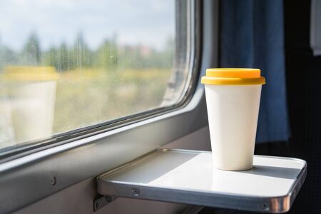 A bamboo coffee cup with a yellow silicon lid. Coffee to go on a table in the train overlooking a beautiful rural green landscape. Travel, lifestyle. Concept zero waste, reusable recyclable dishesの写真素材