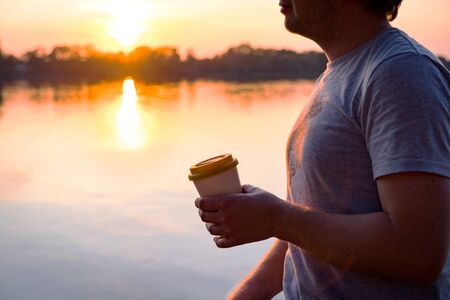 Man is walking in the park near the river and holding a bamboo cup silicone lid with hot coffee in hand. Summer evening rest. Beautiful sunset at the pondの写真素材