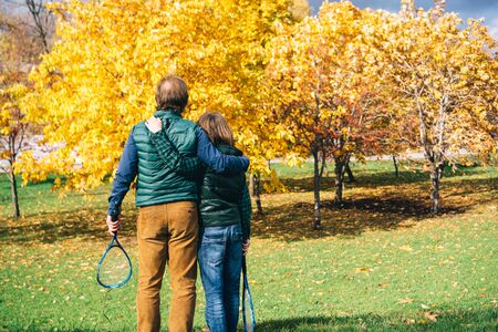 A father and son of school age with badminton racket are looking at sky and talking. Autumn landscape in yellow tones, warm vests and shirts. Sunny funny day. Parenthood and childhood conceptの写真素材