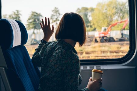 A middle aged woman sitting by the window of a commuter train with coffee in a reusable bamboo cup waving her hand. The concept of remote work, business trips, freelancer, Tourist solo travelの写真素材