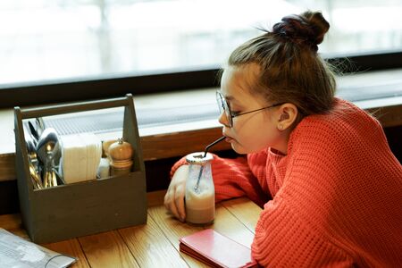 Girl teenager sitting in cafe at a table in red knitted sweater waiting and drinking cocktail through a straw. Milk chocolate cocktail in a glass reusable bottle with a foil cap and a disposable tubeの写真素材