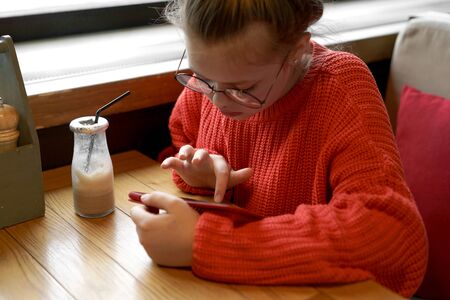 Girl teenager sitting in cafe at a table in red knitted sweater waiting and looking typing in phone messages. Milk chocolate cocktail in a glass reusable bottle with a foil cap and a disposable tubeの写真素材