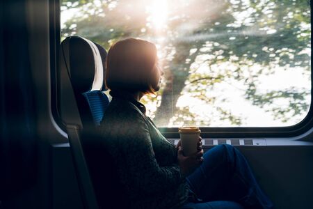A middle aged woman sitting by the window of a commuter train with a laptop and coffee in a reusable bamboo cup in hand. The concept of remote work, business trips, freelancer, Tourist solo travelの写真素材