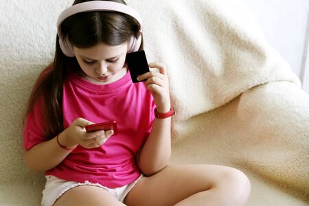 Cheerful girl in pink headphones and a red T-shirt sits with a phone in her hand and a credit card. The concept of gadget and childrens shopping onlineの写真素材