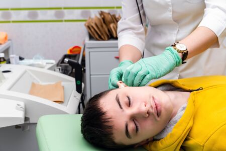 Preventive inspection of a teenage boy in a medical facility. Otorhinolaryngologist doctor pediatrician. Inspection of the sinuses of the ear with the reflector of the forehead and the nasal mirrorの写真素材
