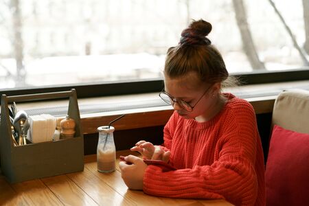 Girl teenager sitting in cafe at a table in red knitted sweater waiting and looking typing in phone messages. Milk chocolate cocktail in a glass reusable bottle with a foil cap and a disposable tubeの写真素材