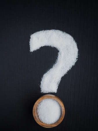 Salt spilled in the shape of question mark and wooden bowl on black background. Pros and cons of table salt.の写真素材