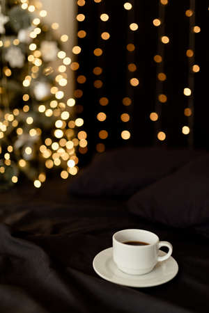 White cup of coffee on a dark bed with pillows in a cozy room with a christmas tree and blurred lights.の写真素材