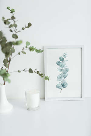 A branch of eucalyptus stands in a vase on a table. On the table a watercolor drawing in a frameの写真素材