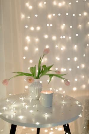 A white vase in pink tulips stands on the table, a candle and New Year's garlands are burning nearby. Blurred lights.の写真素材