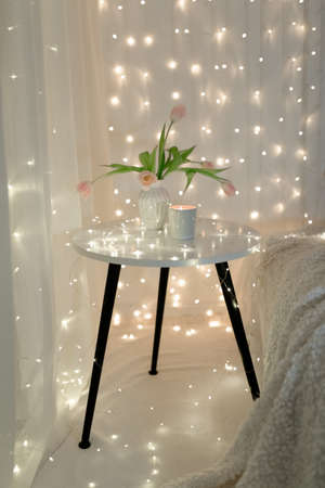 There is a vase in pink tulips on a white table, a candle and New Year's garlands are burning...